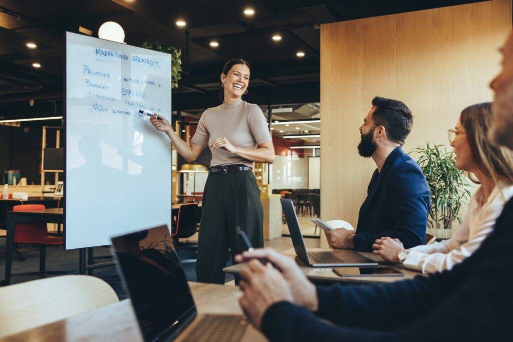 Cheerful business manager giving a board presentation in a modern workspace. Happy young businesswoman smiling while presenting her marketing strategy and ideas to her colleagues.